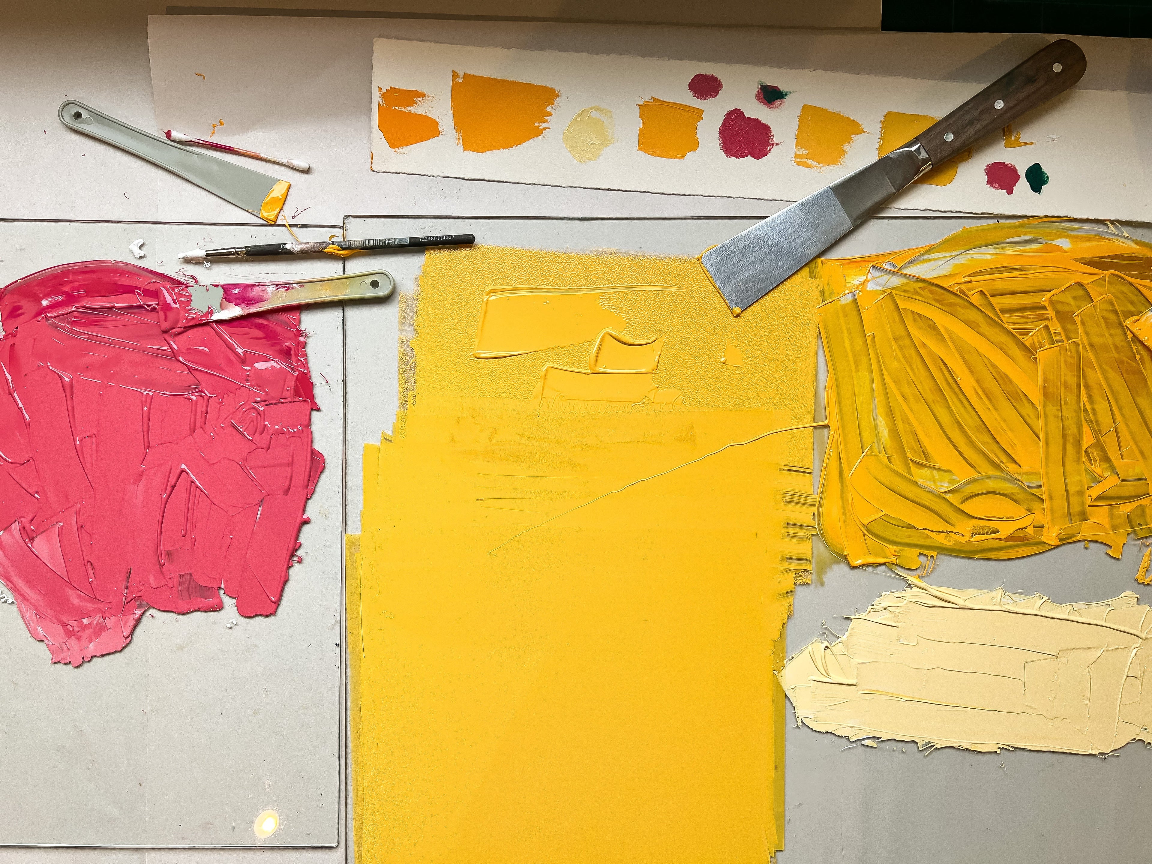An abstract painting with thick, textured brushstrokes of bright yellow, hot pink, and cream on a white canvas, accompanied by artist tools including paint knives and a palette knife.